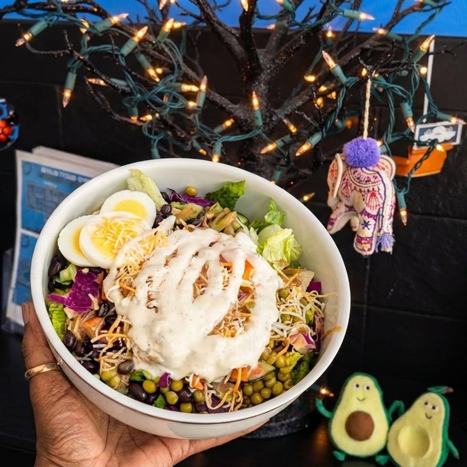 A hand holds a colorful salad bowl with dressing, eggs, and beans, near a lit decorative tree.